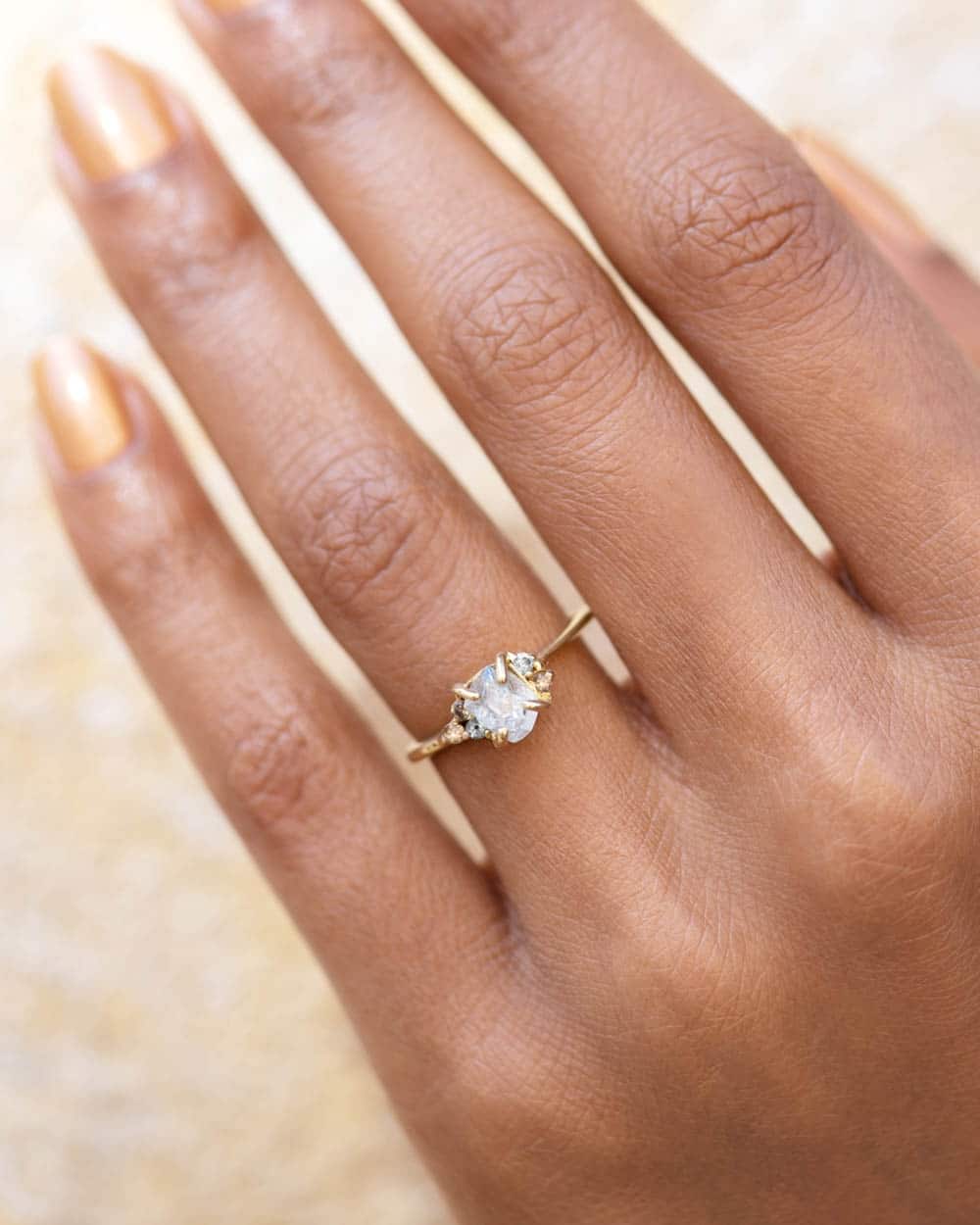 A han displays a gold ring that eatures a central clear uncut diamond surrounded by smaller gemstones, resting against a neutral background.