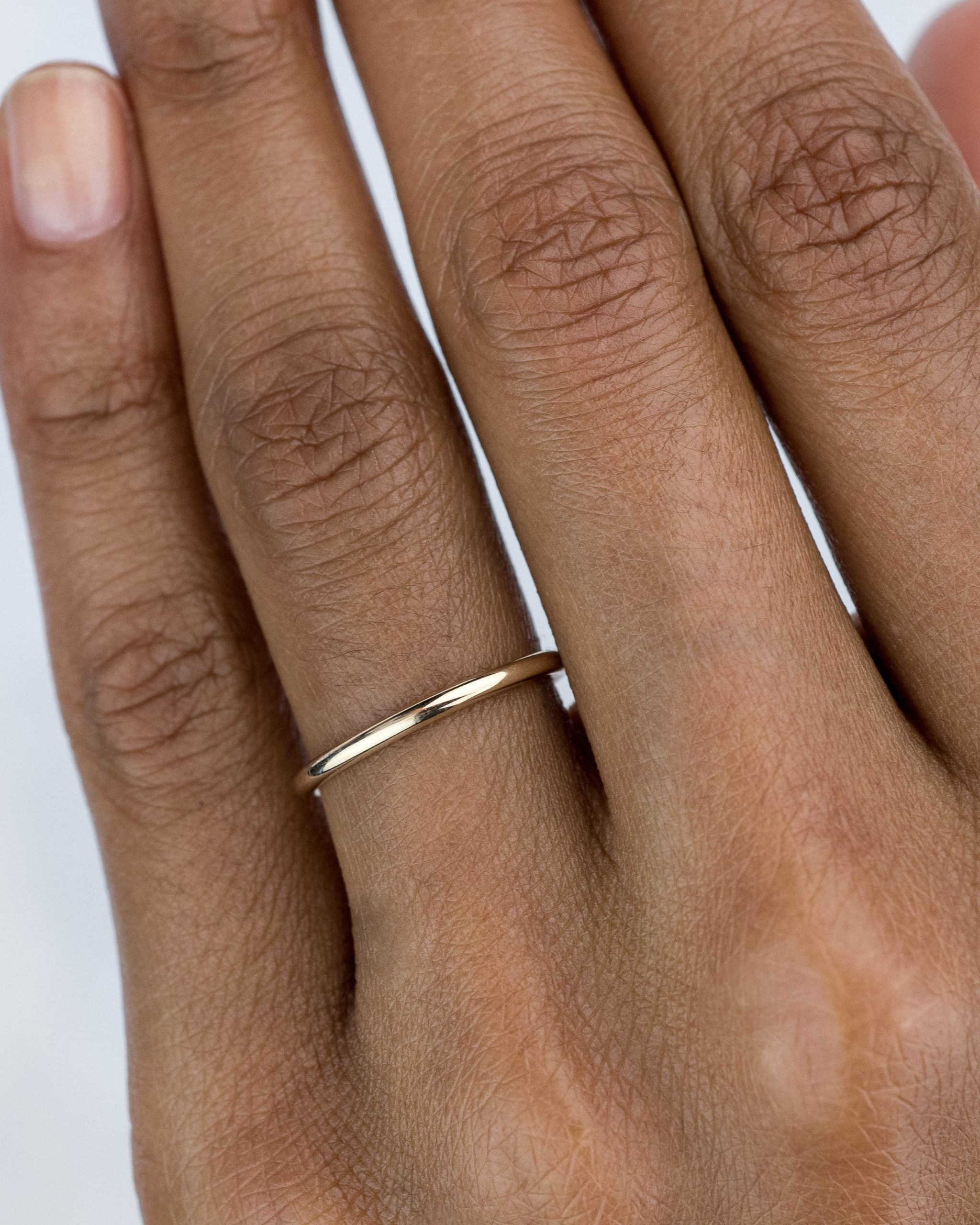 A hand is shown wearing a simple gold ring on the fourth finger. The skin tone is brown, and the background is white, emphasizing the hand and the ring.