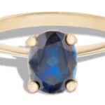 A gold ring features an oval, faceted blue sapphire gemstone as its centerpiece, resting in four prongs. The ring is set against a neutral background, highlighting its sparkling detail.