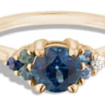 A gold ring features a central dark blue sapphire gemstone flanked by smaller blue and clear stones, showcasing an elegant design meant for adornment, likely in a display setting.