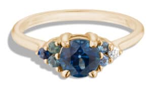 A gold ring features a central dark blue sapphire gemstone flanked by smaller blue and clear stones, showcasing an elegant design meant for adornment, likely in a display setting.