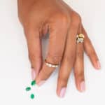 A hand with a stack of several rings is delicately placing three green gemstones on a white surface, showcasing the contrast between the colorful stones and the simple background.