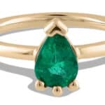 A solid gold ring features a pear-shaped emerald at the center, flanked by small rounded embellishments, reflecting elegance in design, suitable for casual or formal wear.