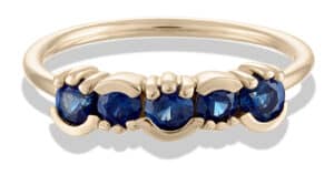 A solid gold ring features five blue sapphires set in a wavy design. The gemstone ring is smooth and polished, standing out against a neutral background, highlighting the vibrant blue stones.