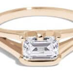 A gold ring features a prominent emerald-cut diamond centerpiece. The ring has a sleek design with an open band, showcasing a modern and elegant aesthetic, ideal for special occasions.