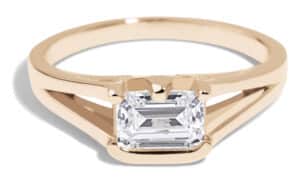 A gold ring features a prominent emerald-cut diamond centerpiece. The ring has a sleek design with an open band, showcasing a modern and elegant aesthetic, ideal for special occasions.
