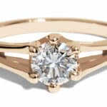 A gold engagement ring features a prominent round white sapphire at the center, supported by a sleek band with a polished finish, placed against a white background for contrast.