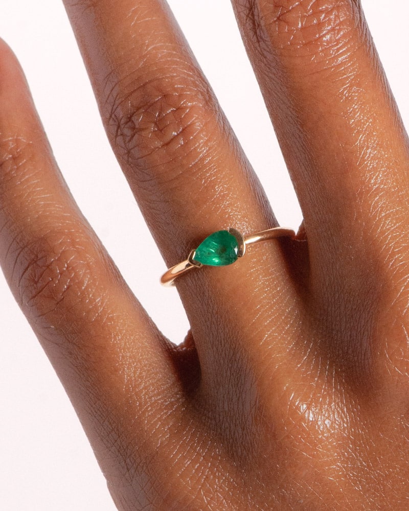 A slender hand with a deep brown skin tone showcases a gold ring featuring an east-west set pear-shaped green gemstone. The background is plain white, emphasizing the jewelry's elegance.