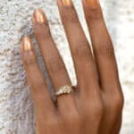 A hand with a yellow gold wedding ring set, featuring multiple sparkling stones, is positioned against a textured, light-colored wall, highlighting the ring's elegance.