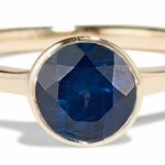 A gold ring features a round, faceted blue gemstone as the centerpiece, reflecting light. It rests on a smooth surface, highlighting its simple yet elegant design.
