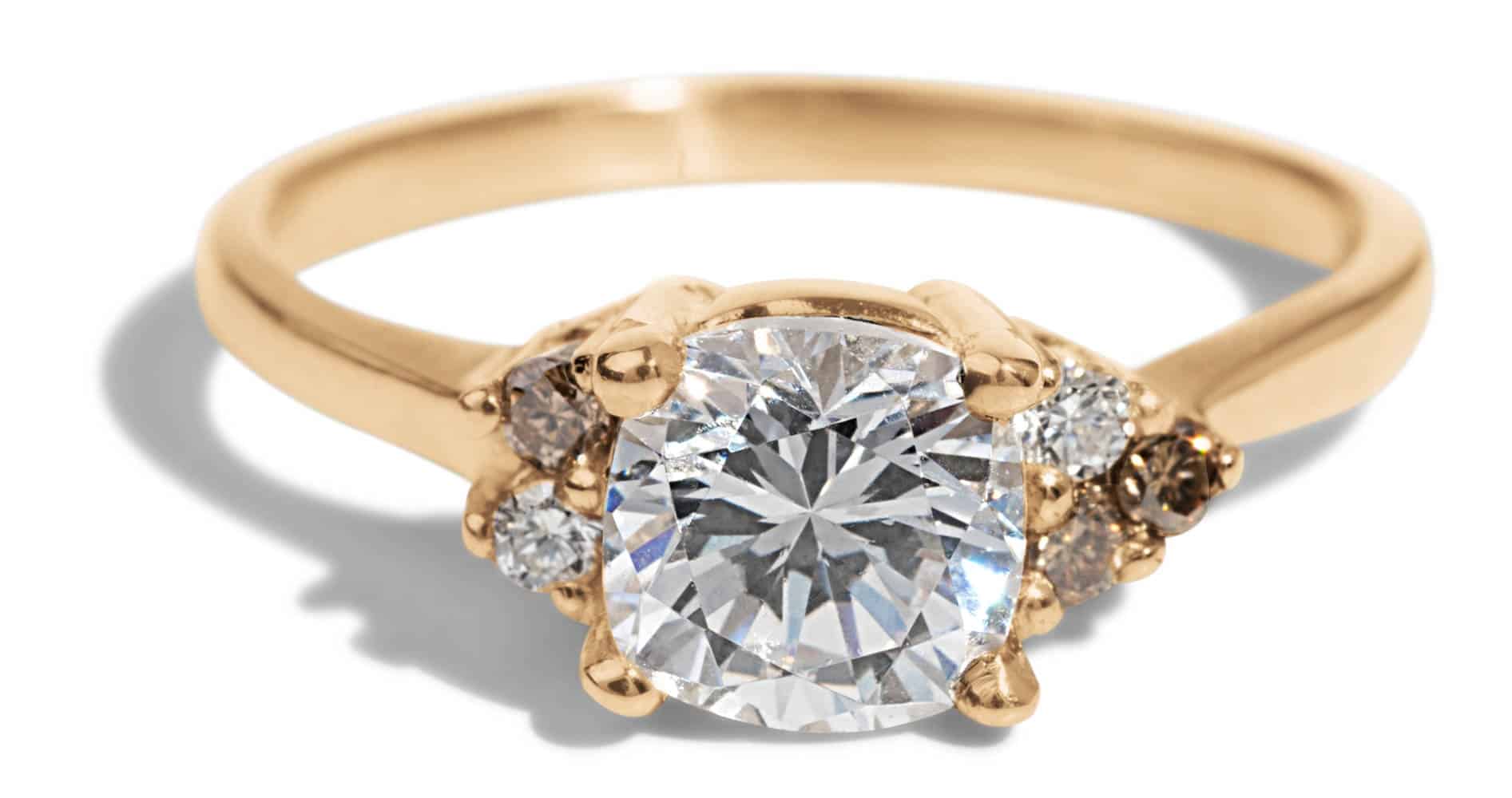 A gold engagement ring features a large, radiant central diamond, flanked by smaller diamonds, glimmering against a soft, neutral background. The design highlights elegance and craftsmanship.