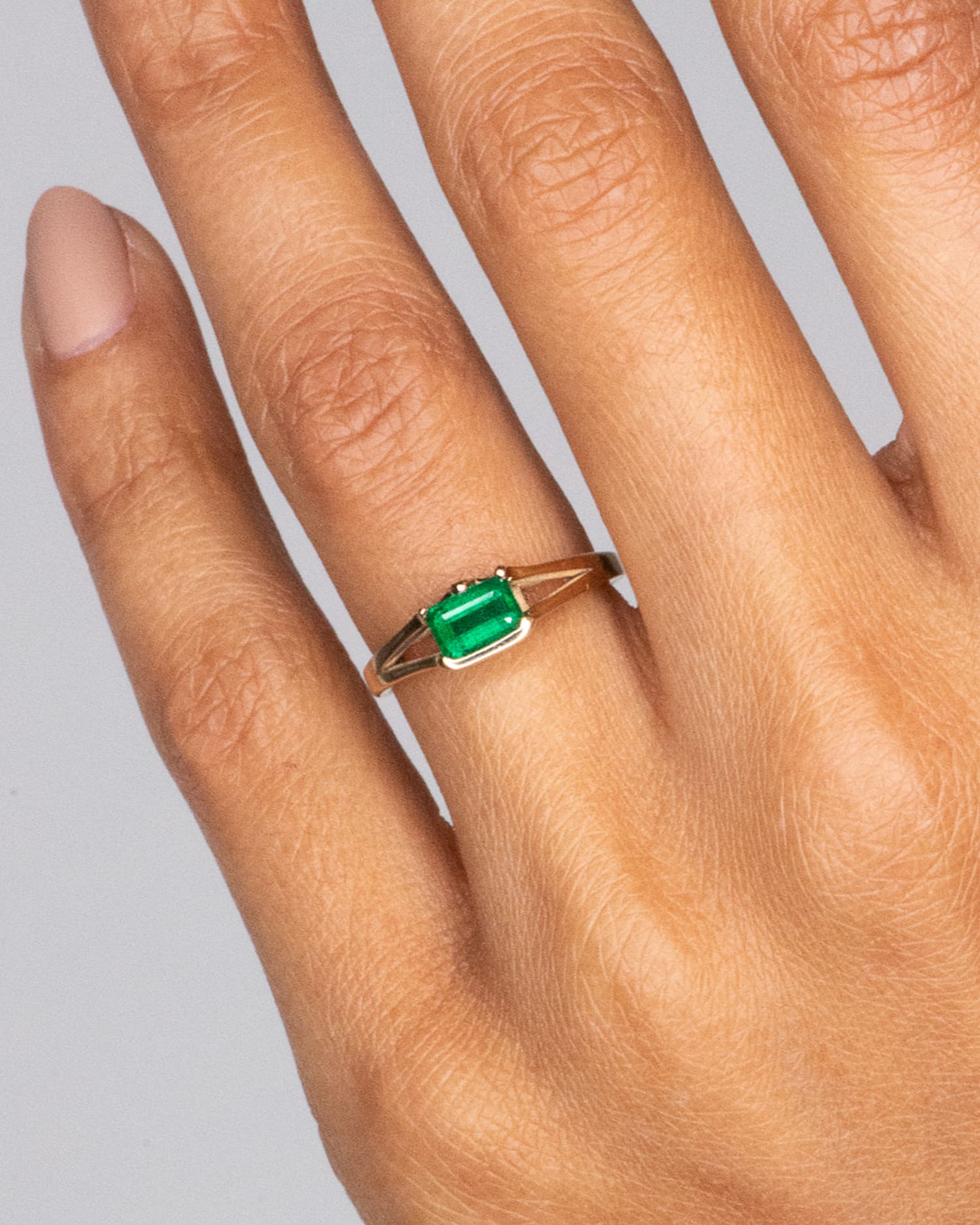 A hand with a softly manicured nail showcases a delicate gold ring featuring a prominent rectangular green emerald.