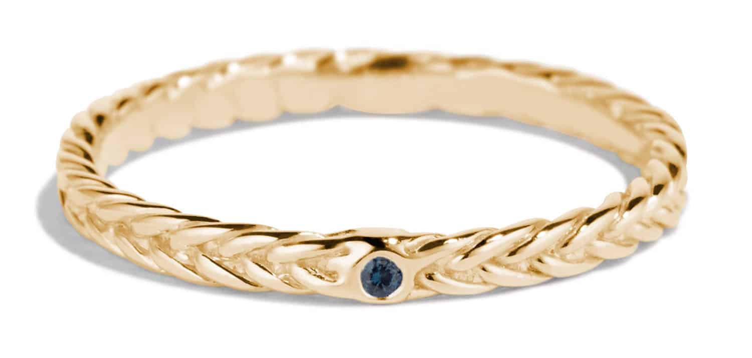 A gold braided ring featuring a small blue sapphire gemstone is displayed gracefully, highlighting its intricate design against a neutral background. The ring’s shape is circular and delicate.