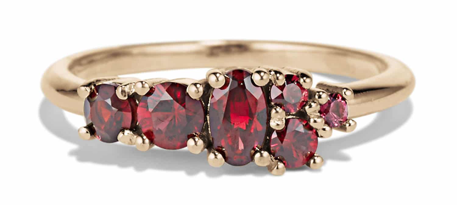 A gold ring features five clustered red gemstones, including oval and round cuts, set in a delicate bezel. The background is plain, emphasizing the ring's design.