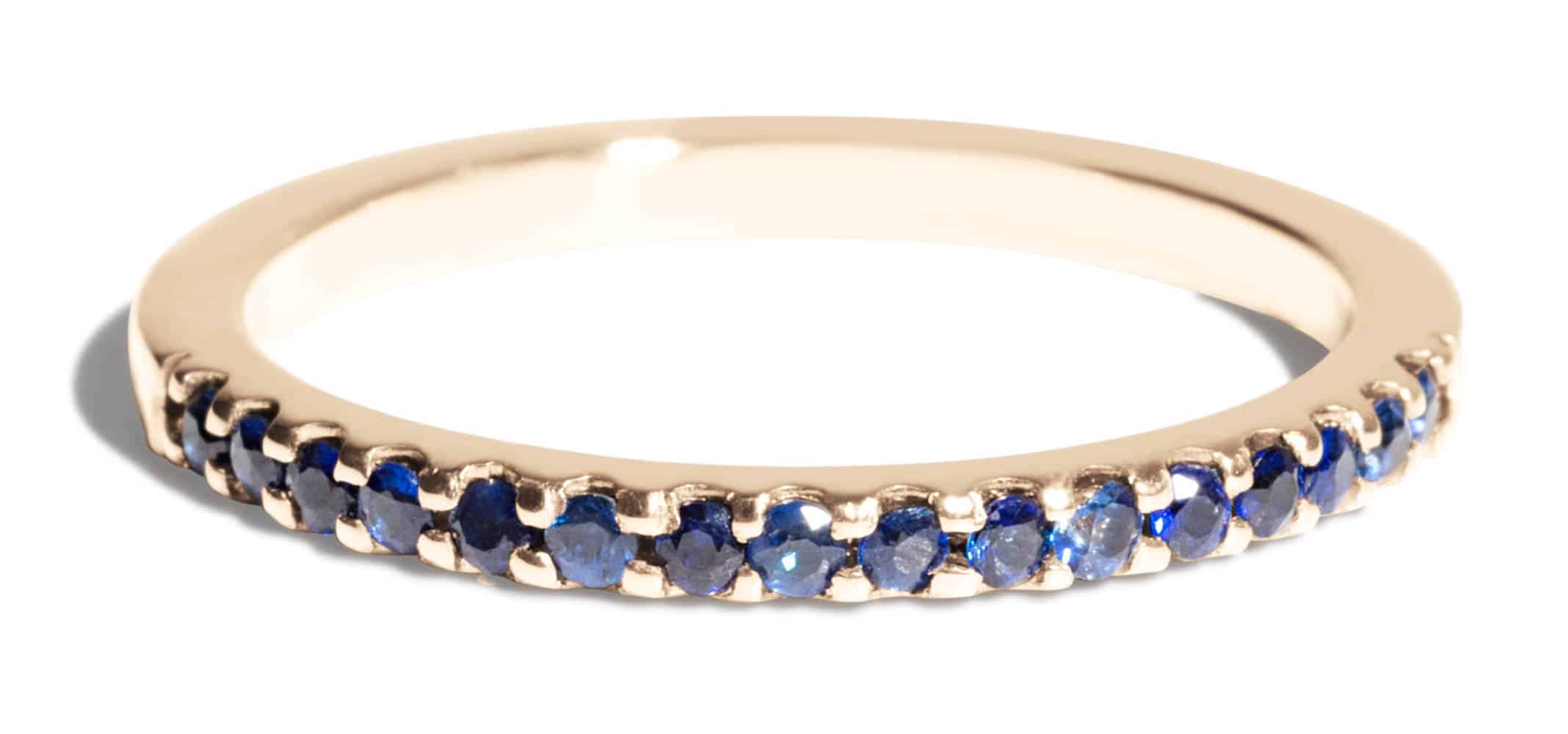 A slender wedding band ring features a row of blue gemstones embedded along its surface, set against a smooth, gold-colored metal. The setting is simple and elegant, highlighting the stones.