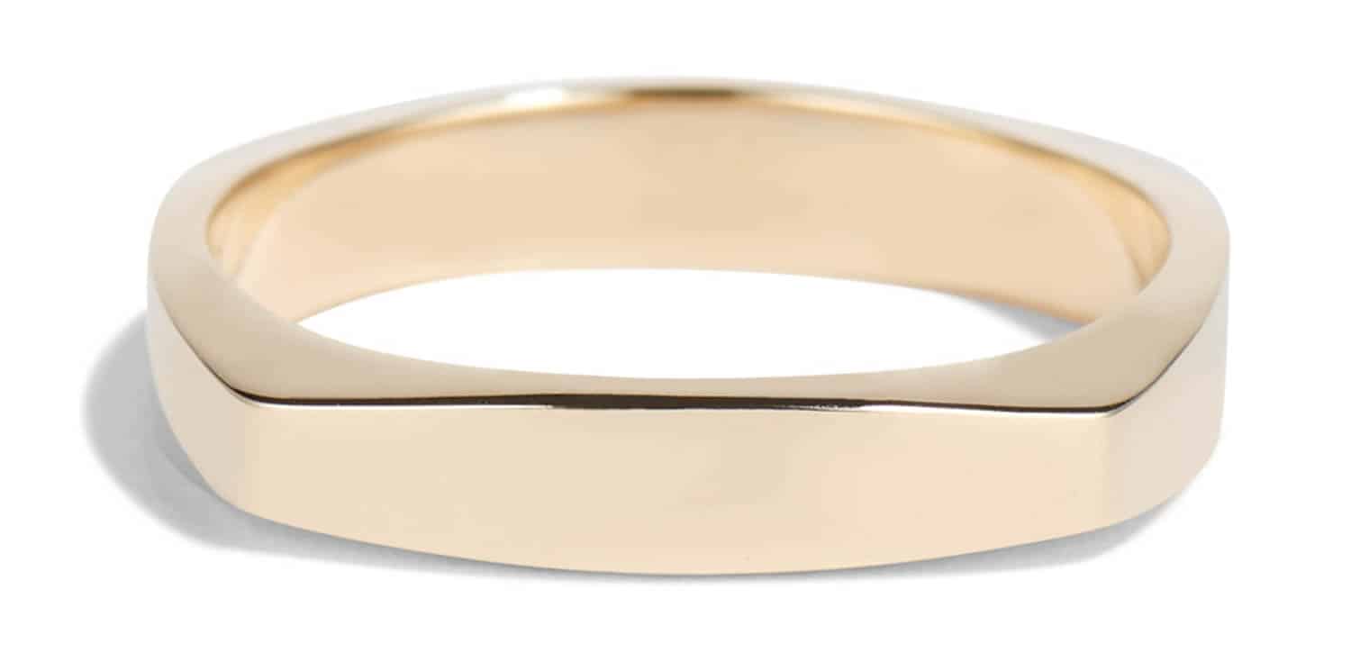 A smooth, wide gold band lies horizontally, showcasing its polished surface and angular edges, reflecting soft light against a minimalistic white backdrop, emphasizing its elegant design.