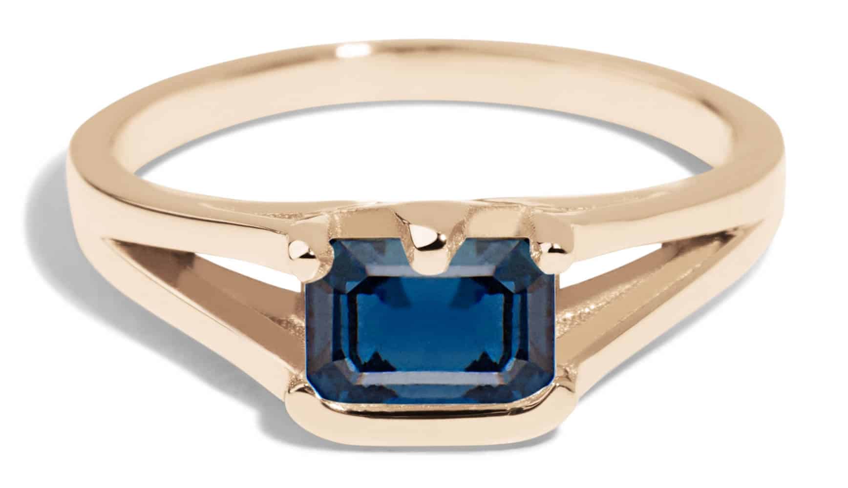 A gold ring features a rectangular blue sapphire gemstone as its centerpiece, set in a simple yet elegant design, highlighting the stone against a neutral background.