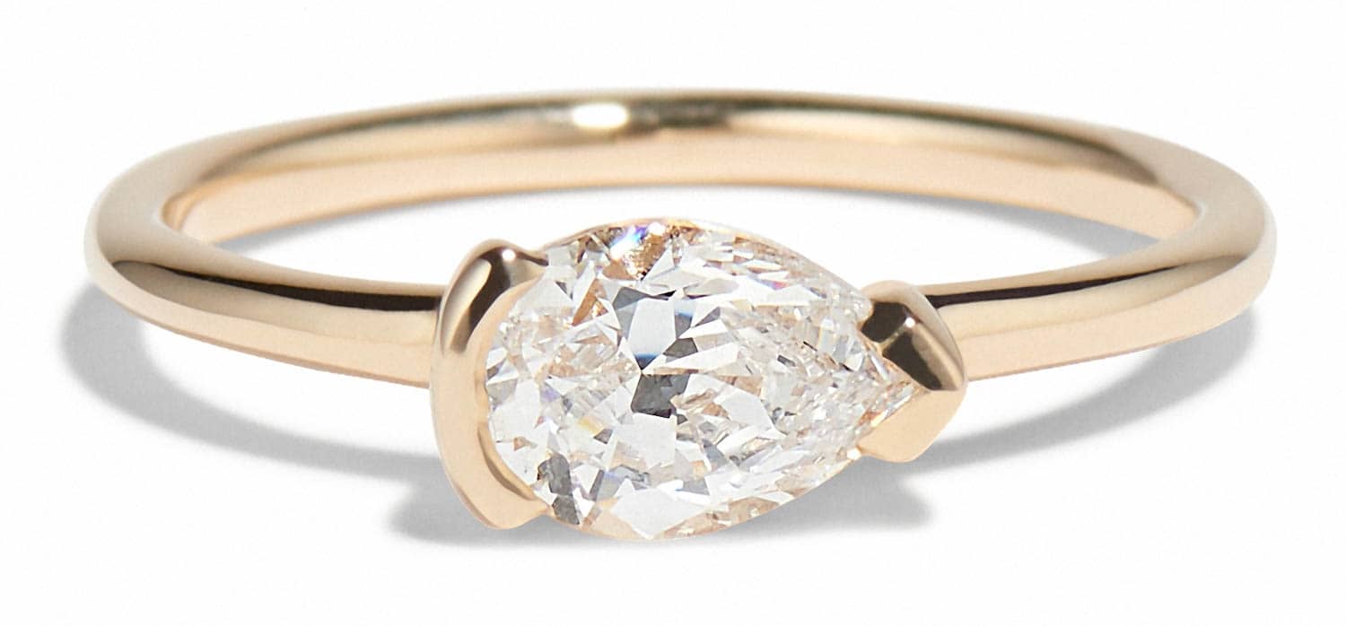 A gold ring features an east- west set pear-shaped diamond at its center, elegantly set and glimmering, resting against a plain white background that highlights its shine and detail.