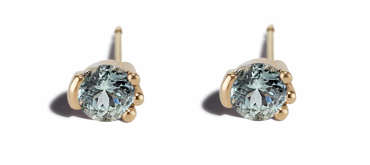 Two elegant gold stud earrings feature large, round, light blue sapphire gemstones. They are positioned symmetrically against a clean, white background, highlighting their sparkle and refined design.