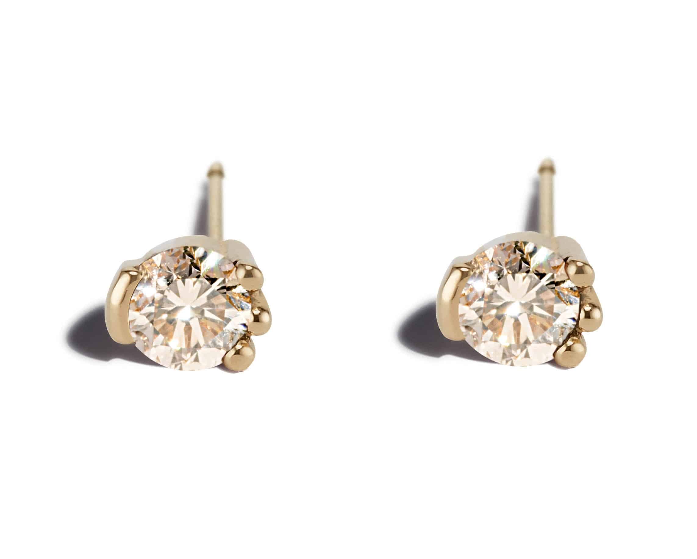 Two golden diamond stud earrings are displayed. Each features a round, sparkling diamond held by four prongs. The background is plain white, emphasizing the jewelry's shine and elegance.