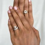 A close-up view of two hands, one adorned with a sparkling diamond ring on the ring finger, and the other hand positioned on top, showcasing another exquisite diamond ring.
