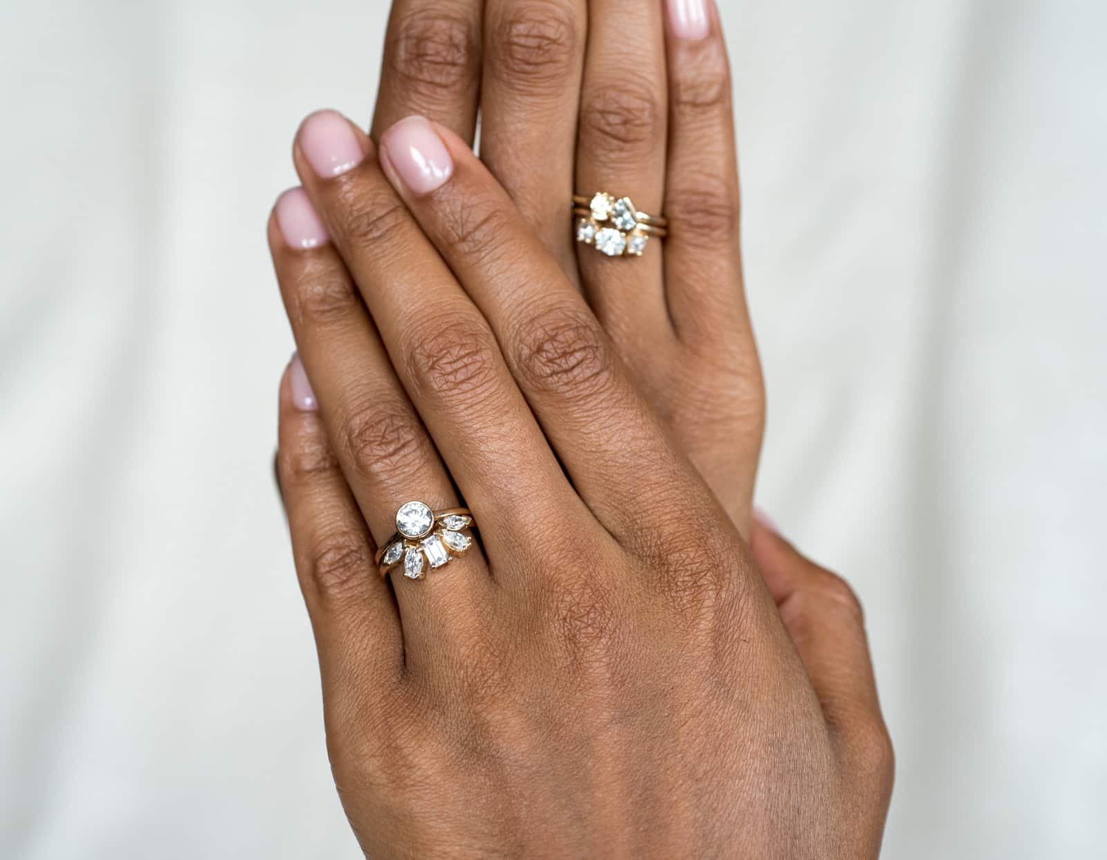 A close-up view of two hands, one adorned with a sparkling diamond ring on the ring finger, and the other hand positioned on top, showcasing another exquisite diamond ring.