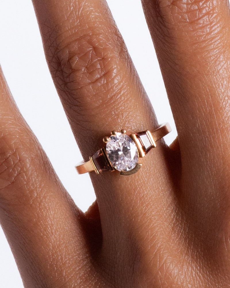 A hand wearing a ring with an oval diamond in a gold semi-bezel setting, flanked by two smaller rectangular stones, against a plain white background.
