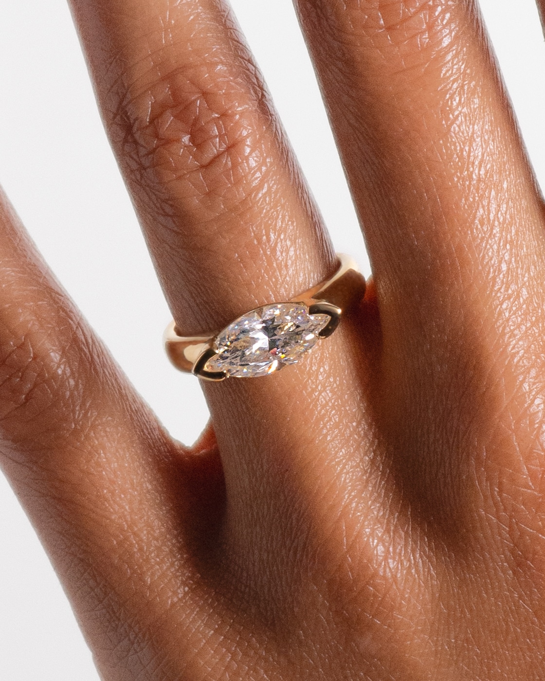 A gold ring with a marquise diamond is prominently displayed on a hand, showcasing the ring's sparkle against the warm skin tone and intricate finger details.